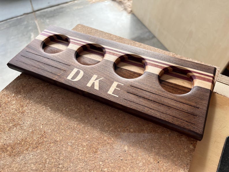 May include: A rectangular wooden beer flight paddle featuring four circular cutouts for glasses. The board is crafted from dark brown wood with light and purple-toned wood accents. The letters DKE are engraved in the center with horizontal lines.
