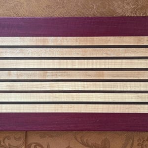 May include: A rectangular wooden cutting board with a striped design. The board features alternating light-colored wood stripes with dark brown and black accents, and a deep purple border at the top and bottom. The cutting board is approximately 18 inches long and 12 inches wide.