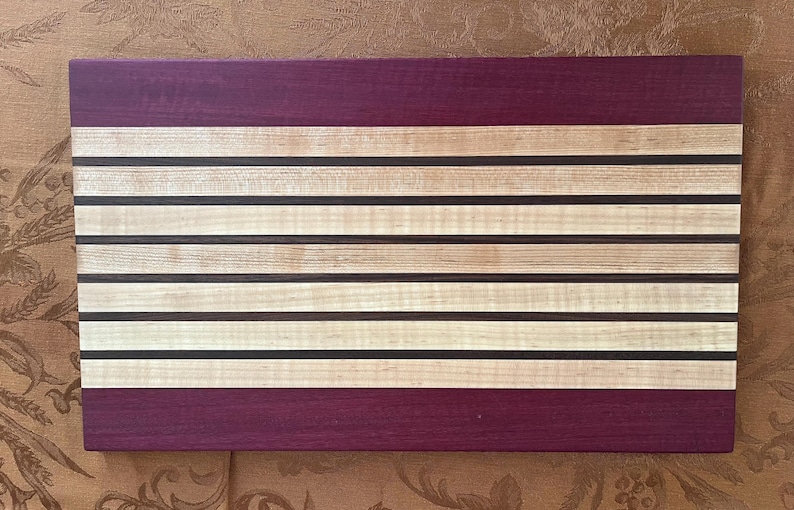 May include: A rectangular wooden cutting board with a striped design. The board features alternating light-colored wood stripes with dark brown and black accents, and a deep purple border at the top and bottom. The cutting board is approximately 18 inches long and 12 inches wide.