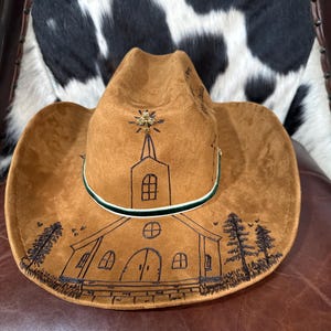 May include: Brown cowboy hat with a church illustration. The hat features a gold-colored embellishment at the crown and a green and white band around the base of the crown. The church design is in black ink.