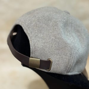 May include: A gray baseball cap with a brown leather strap and a brass buckle. The cap is made of a soft, felt-like material. The strap is adjustable.