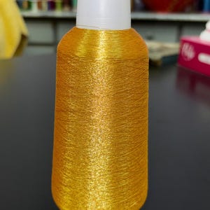 Può includere: Una bobina di filo dorato metallizzato con base e cappuccio bianchi. Il filo è avvolto strettamente, creando una superficie strutturata. Ideale per ricamo, cucito o altri progetti creativi.