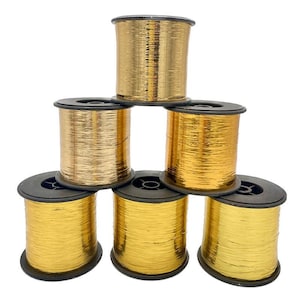 Può includere: Una piramide di sei rocchetti di filo dorato metallico. Ogni rocchetto ha una base e un coperchio in plastica nera. Il filo appare lucido e riflettente, suggerendo un materiale metallico. I rocchetti sono su uno sfondo bianco.