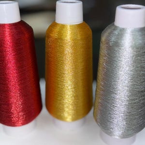 Può includere: Tre rocchetti di filo metallizzato rosso, oro e argento. Ogni rocchetto ha una base e una parte superiore bianche. Il filo ha una qualità lucida e riflettente, suggerendo che sia fatto di un materiale sintetico.