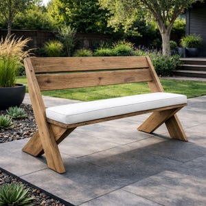 May include: A wooden outdoor bench with a white cushion. The bench has a slatted back and angled legs, constructed from light brown wood. The white cushion provides a contrast to the wood. The bench is placed on a gray stone patio.