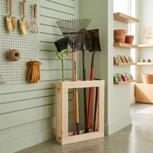May include: A light-colored wooden garden tool organizer rack holding a metal rake, a shovel, a spade, and a green broom. The rack is rectangular with a sturdy frame, designed to keep long-handled gardening implements upright and organized.