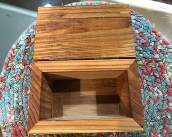 May include: A rectangular wooden box with a hinged lid, featuring a natural light brown wood grain finish. The box is open, revealing a hollow interior with light-coloured wood panels and a visible metal hinge connecting the lid to the base.