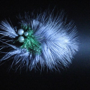 Könnte beinhalten: Dekorative Haarspange mit weißen und blauen Federn, verziert mit drei Kunstperlen und einem Hauch von Grün. Der Clip ist vor einem dunklen Hintergrund platziert, der die zarten Texturen und Farben hervorhebt.