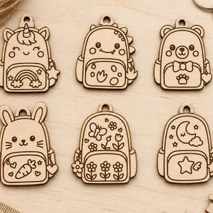 May include: Six laser-cut wooden backpack-shaped charms featuring engraved designs of a unicorn with a rainbow, a dinosaur, a bear, a rabbit with a carrot, a butterfly with flowers, and a moon with a star, each with a top hole for hanging.