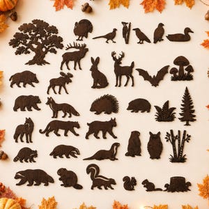 Pode incluir: Uma coleção de recortes de madeira castanho-escuro com vários animais da floresta, aves, árvores e plantas, incluindo um veado, urso, raposa, coruja e esquilo, dispostos numa superfície clara rodeada por folhas de outono e pinhas.