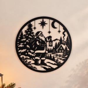 Op de afbeelding: Een cirkelvormige wanddecoratie van zwart metaal met een silhouet van de Kerstman die een lantaarn vasthoudt in een besneeuwd bos. De afbeelding bevat dennenbomen, een huisje met rook uit de schoorsteen, een maansikkel en sterren.