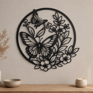 Peut inclure: Une décoration murale circulaire noire présentant un motif découpé de deux papillons entourés de branches fleuries et de feuilles. La silhouette métallique complexe affiche un grand papillon au centre et un plus petit près du bord supérieur.