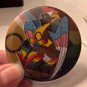 Op de afbeelding: Een ronde knoop met een kleurrijke illustratie van een mythisch wezen met regenboogaccenten. Het wezen heeft vleugels en is gehuld in een regenboog gestreept ontwerp. De knoop wordt door een hand vastgehouden.