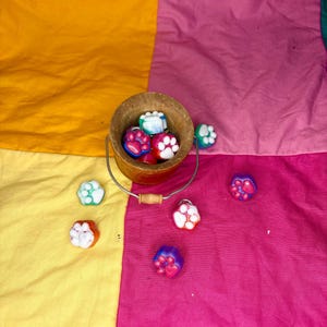 May include: A wooden bucket with a metal handle is filled with colorful paw-shaped items. Several paw prints are scattered on a patchwork background of yellow, pink, and orange fabric. The paw prints are in shades of blue, pink, and white.