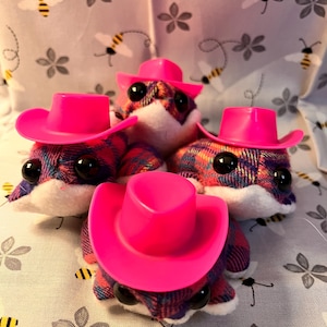 May include: Four plush toy creatures with plaid patterned bodies and white faces, each wearing a bright pink cowboy hat. The toys have large black eyes and are arranged on a fabric with a bee and flower pattern.