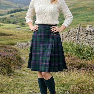Puede incluir: Una falda tipo kilt plisada hasta la rodilla con un patrón de cuadros en verde oscuro, azul y líneas rojas finas. La falda incluye una correa lateral de cuero con una hebilla de metal plateado en la cintura, combinada con un suéter de punto trenzado.