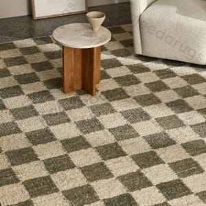 Puede incluir: Una alfombra a cuadros de tonos neutros con un patrón repetitivo de cuadrados grises oscuros y crema. Una pequeña mesa auxiliar redonda con tapa de mármol y base de madera se encuentra sobre la alfombra, con un jarrón decorativo encima. Un sillón blanco está en el fondo.