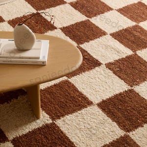 May include: A brown and cream checkered rug with a round wooden coffee table. A small, off-white vase with a sprig of berries sits on top of a stack of books. The rug's pattern is a repeating grid of squares.