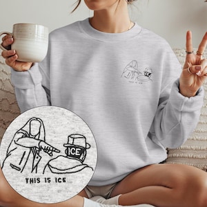 May include: A light gray crewneck sweatshirt featuring a small black embroidered line art design on the left chest. The graphic depicts one figure pointing at another wearing a hat with the text "THIS IS ICE" embroidered below the figures.