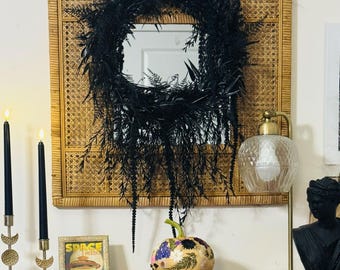 May include: A black wreath hangs over a square mirror set in a woven rattan frame. Below, a decorative pumpkin with floral designs sits atop books. Two black candles stand in gold candle holders, and a gold lamp with a glass shade is on the right.