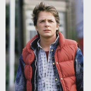 Puede incluir: Un hombre con un chaleco acolchado rojo sobre una chaqueta vaquera y una camisa a cuadros. El hombre tiene el pelo castaño claro y mira a la cámara. La imagen es un plano medio, con un fondo neutro.