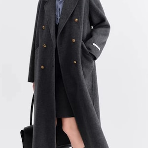 Könnte beinhalten: Ein dunkelgrauer, doppelreihiger Mantel mit goldenen Knöpfen. Der lange Mantel hat ein klassisches Design mit einem Reverskragen und Vordertaschen. Das Model trägt ein schwarzes Kleid, graue Socken und schwarze Schuhe. Eine schwarze Tragetasche wird in der linken Hand gehalten.