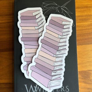 Puede incluir: Dos pegatinas de libros apilados en tonos rosas y morados descansan sobre un libro negro. Las pegatinas tienen un borde blanco. El libro negro tiene el nombre "Winters" y el texto "USA TODAY BESTSELLING AUTHOR" impreso.