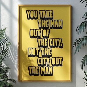 May include: A framed poster with the quote "YOU TAKE THE MAN OUT OF THE CITY, NOT THE CITY OUT THE MAN" in black text on a yellow background. The poster is in a gold frame and is surrounded by green plants.