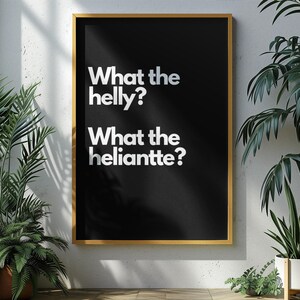Puede incluir: Un cuadro negro enmarcado con borde dorado, con las frases blancas "What the helly?" y "What the heliantte?". La obra de arte está colgada en una pared blanca, rodeada de plantas en macetas.