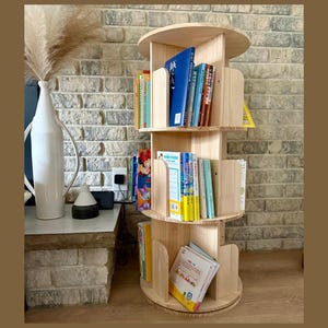 May include: A natural wood, rotating bookshelf with multiple circular shelves. The shelves are filled with colorful books of various sizes. The bookshelf is made of light-colored wood and is placed against a brick wall.
