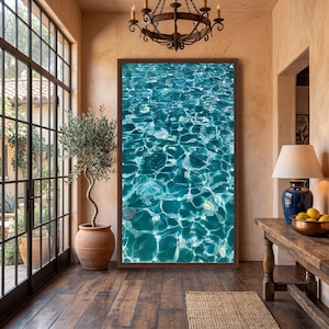 May include: A large rectangular wall art piece featuring a high-resolution photograph of shimmering turquoise swimming pool water with bright white light reflections and ripples, framed in a dark brown wood frame hanging on a beige wall.