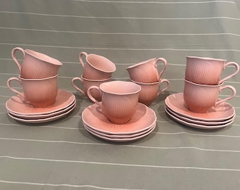 May include: A set of eight pink teacups and saucers. The cups and saucers are a soft pink with a ribbed design. Each saucer has a white trim. The cups are stacked in pairs, and the saucers are stacked in sets of three.