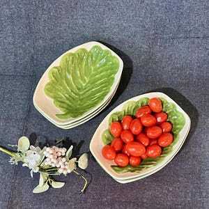 May include: Two leaf-shaped ceramic serving dishes, one filled with red cherry tomatoes. The dishes have a green leaf design on a cream background. A small floral arrangement is in the lower left corner.