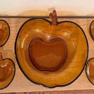 May include: A set of amber-colored glass serving dishes in the shape of apples. The set includes a large bowl and four smaller dishes, all with a black trim. The dishes are arranged on a wooden surface.