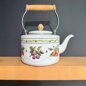 Puede incluir: Una tetera de esmalte blanco con asa y pomo de tapa de madera clara. El cuerpo está decorado con un borde festoneado verde e ilustraciones coloridas de ciruelas, cerezas y bayas, junto con etiquetas de texto de conservas de frutas.