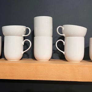 May include: A collection of white ceramic mugs and teacups with a subtle textured pattern. The mugs have curved handles and are arranged on a wooden shelf. The teacups are stacked on top of the mugs.