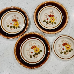 May include: Four-piece vintage dinnerware set. Each piece features a cream-colored center with a floral design in red, yellow, and white. The plates are set on a dark brown rim with a textured edge, creating a warm, inviting aesthetic.