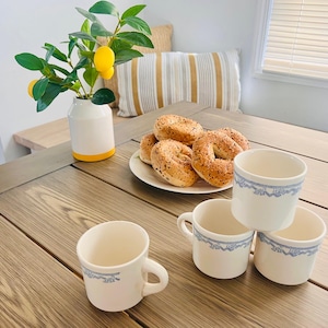 May include: A wooden table setting featuring white mugs with blue floral trim, a plate of bagels, and a vase with lemon branches. The mugs are stacked and arranged on the table.