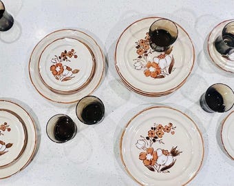 May include: A vintage cream-colored dinnerware set with a floral design in shades of orange and brown. The set includes plates of various sizes and small, dark-colored glasses. The plates have a brown trim.