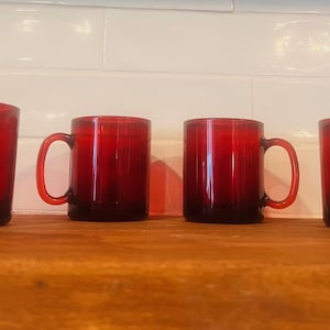 Op de afbeelding: Vier robijnrode glazen mokken met afgeronde handvatten staan op een houten oppervlak. De mokken hebben een eenvoudige, cilindrische vorm en een dieprode kleur, met een iets donkerdere basis. Ze zijn gelijkmatig verdeeld.