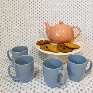 Pode incluir: Uma chaleira rosa claro salpicada com uma bandeja de biscoitos e quatro canecas azul claro. As canecas estão dispostas em frente à chaleira, criando um conjunto de chá. Os biscoitos são dourados e castanhos claros.