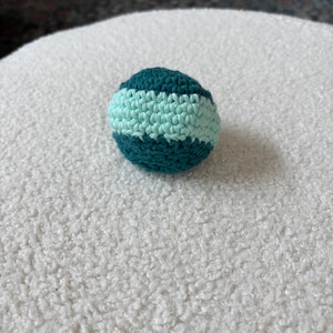 May include: A small, round, crocheted ball with alternating bands of teal and light blue. The ball is handmade and appears to be made of soft yarn. The ball is sitting on a white, textured surface.