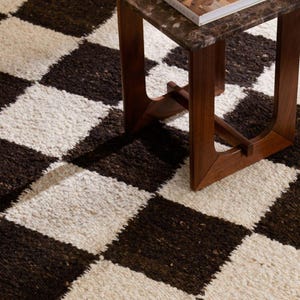 Puede incluir: Primer plano de una alfombra a cuadros marrón y blanca. Una pequeña mesa auxiliar de madera oscura con tapa de mármol y un libro en el fondo. El patrón de la alfombra es una cuadrícula repetida de cuadrados marrón oscuro y blanco roto.