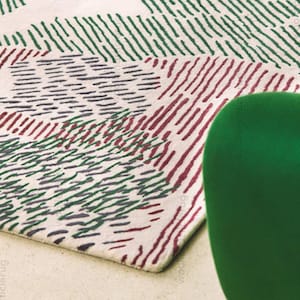 Puede incluir: Una alfombra rectangular con fondo crema y un patrón de líneas verdes, burdeos y azul oscuro. Las líneas están dispuestas en trazos paralelos, creando un efecto texturizado. Un objeto verde está en primer plano.
