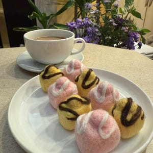 May include: A white teacup and saucer with a beverage, next to a plate of colorful, felted pastries. The pastries are yellow and pink with brown and white accents. Purple flowers and greenery are in the background.