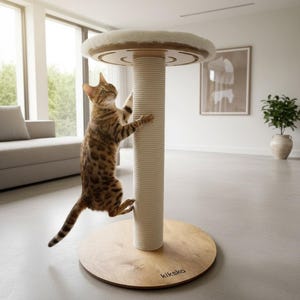 Puede incluir: Un rascador para gatos con base circular de madera que incluye el texto kikska, un pilar central alto envuelto en cuerda color crema y una plataforma superior redonda cubierta de felpa blanca suave, con un gato atigrado marrón trepando.