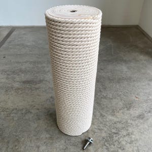 Pode incluir: Um poste arranhador cilíndrico envolto em corda grossa trançada de cor branco-sujo. A superfície superior é de madeira plana de cor clara com um ponto de fixação central de metal. Um pequeno parafuso de metal com arruela está no chão.