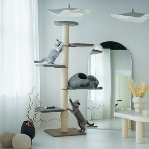 Puede incluir: Un árbol para gatos de varios niveles con múltiples plataformas, un poste rascador y una casa para gatos. Dos gatos grises están jugando en el árbol. El árbol para gatos es beige y gris, con una base cuadrada. El fondo incluye una cortina blanca, un espejo y objetos decorativos.
