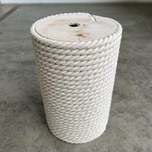Pode incluir: Um banco ou mesa lateral cilíndrica com um tampo de madeira natural clara com veios visíveis e um parafuso central de metal. As laterais externas são envolvidas por uma corda grossa trançada na cor creme, criando um acabamento texturizado.