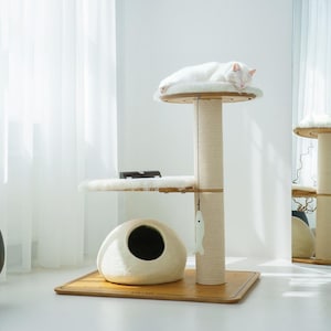 Peut inclure: Un chat blanc se repose sur un arbre à chat à plusieurs niveaux. L'arbre à chat comprend un poteau enveloppé de sisal, une plate-forme ronde avec un coussin doux et blanc et une petite maison pour chat fermée. Un jouet en forme de poisson est suspendu à l'arbre.